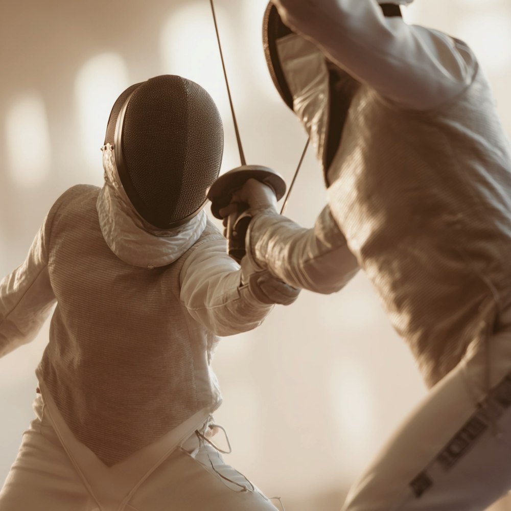 Two fencers in white protective gear mid-bout with crossed swords in a warmly lit training hall