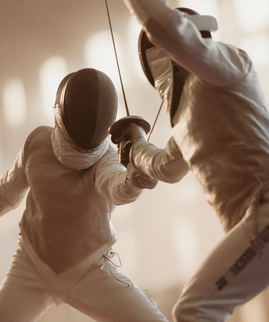 Two fencers in white protective gear mid-bout with crossed swords in a warmly lit training hall