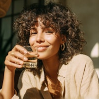 Woman with curly hair smiling while holding a glass of water in warm natural sunlight